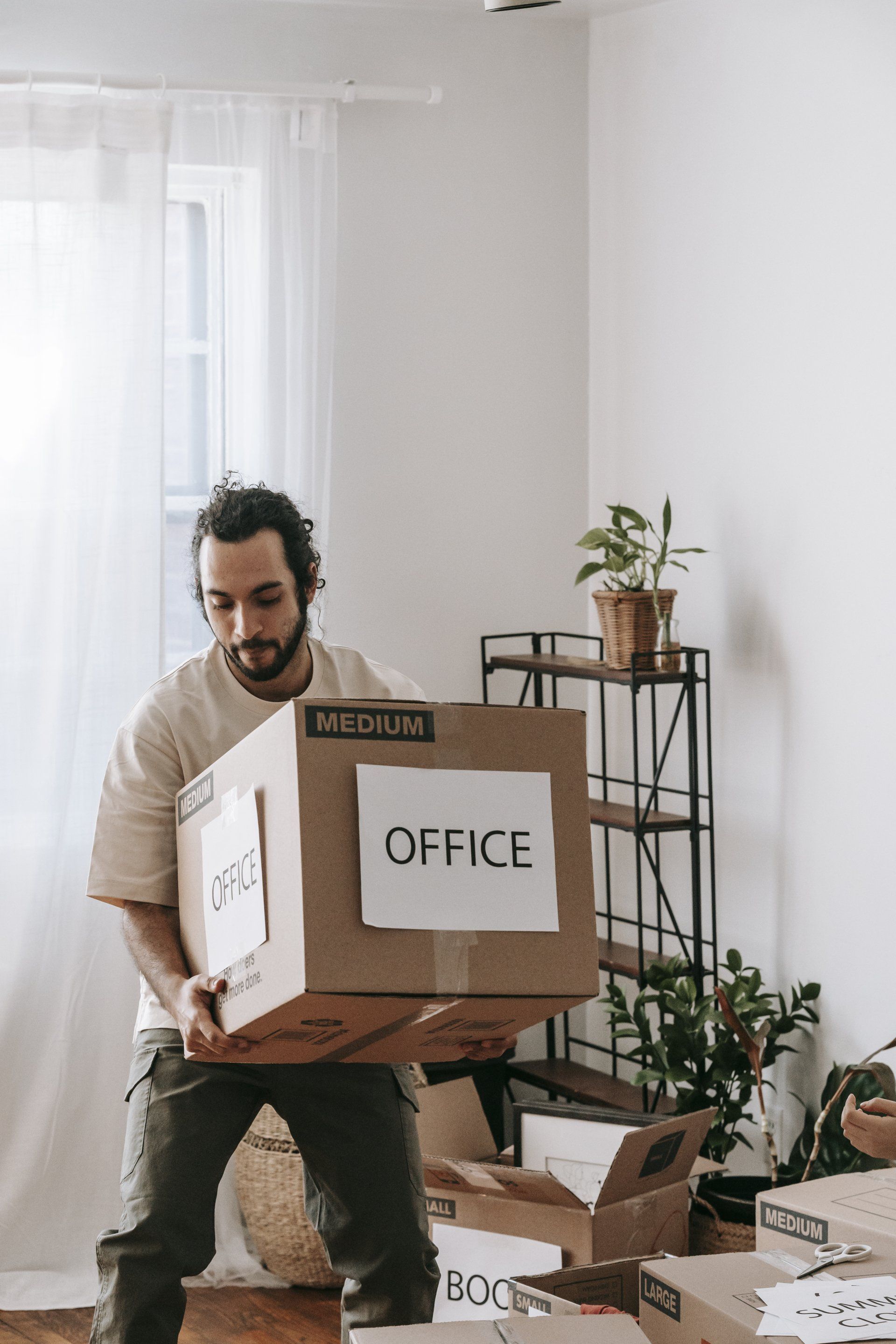Man carrying a cardboard box labeled "OFFICE" during a move. Room interior with other boxes and shelving.