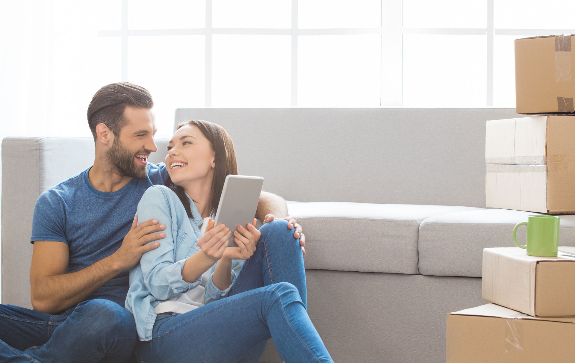 Couple sitting on floor, looking at tablet, smiling, boxes nearby, near a sofa.
