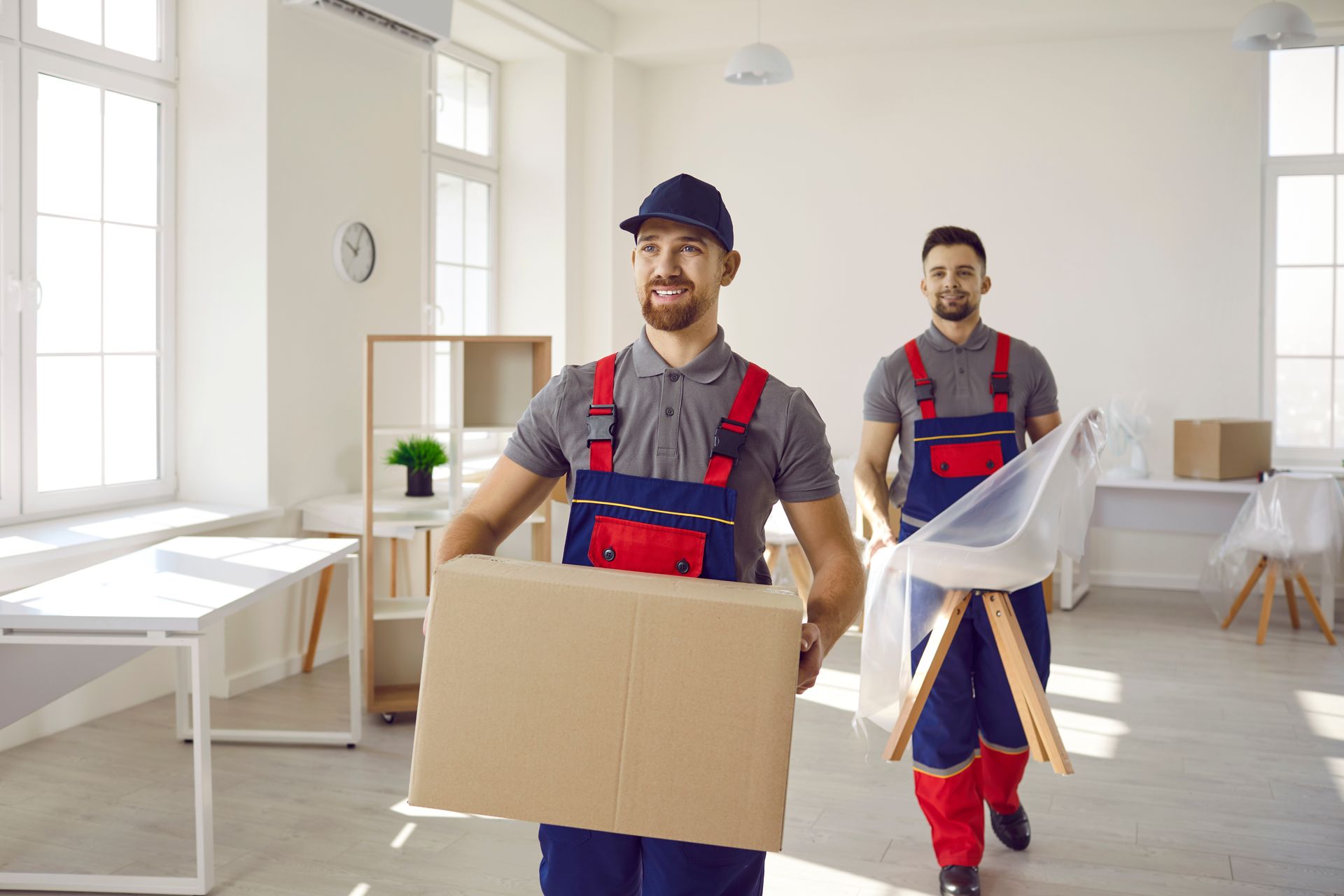 Two movers carry a box and chair in a bright, empty room with desks and windows.